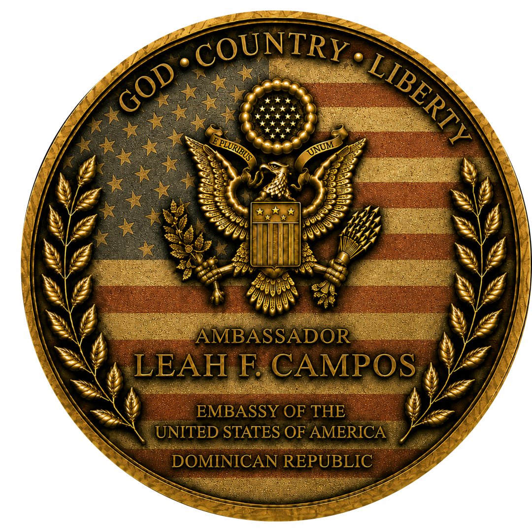 A circular gold-toned emblem or seal featuring an eagle over a distressed American flag design, with laurel branches on both sides and text reading 'GOD • COUNTRY • LIBERTY,' 'AMBASSADOR LEAH F. CAMPOS,' and 'EMBASSY OF THE UNITED STATES OF AMERICA DOMINICAN REPUBLIC.