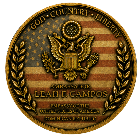 A circular gold-toned emblem or seal featuring an eagle over a distressed American flag design, with laurel branches on both sides and text reading 'GOD • COUNTRY • LIBERTY,' 'AMBASSADOR LEAH F. CAMPOS,' and 'EMBASSY OF THE UNITED STATES OF AMERICA DOMINICAN REPUBLIC.