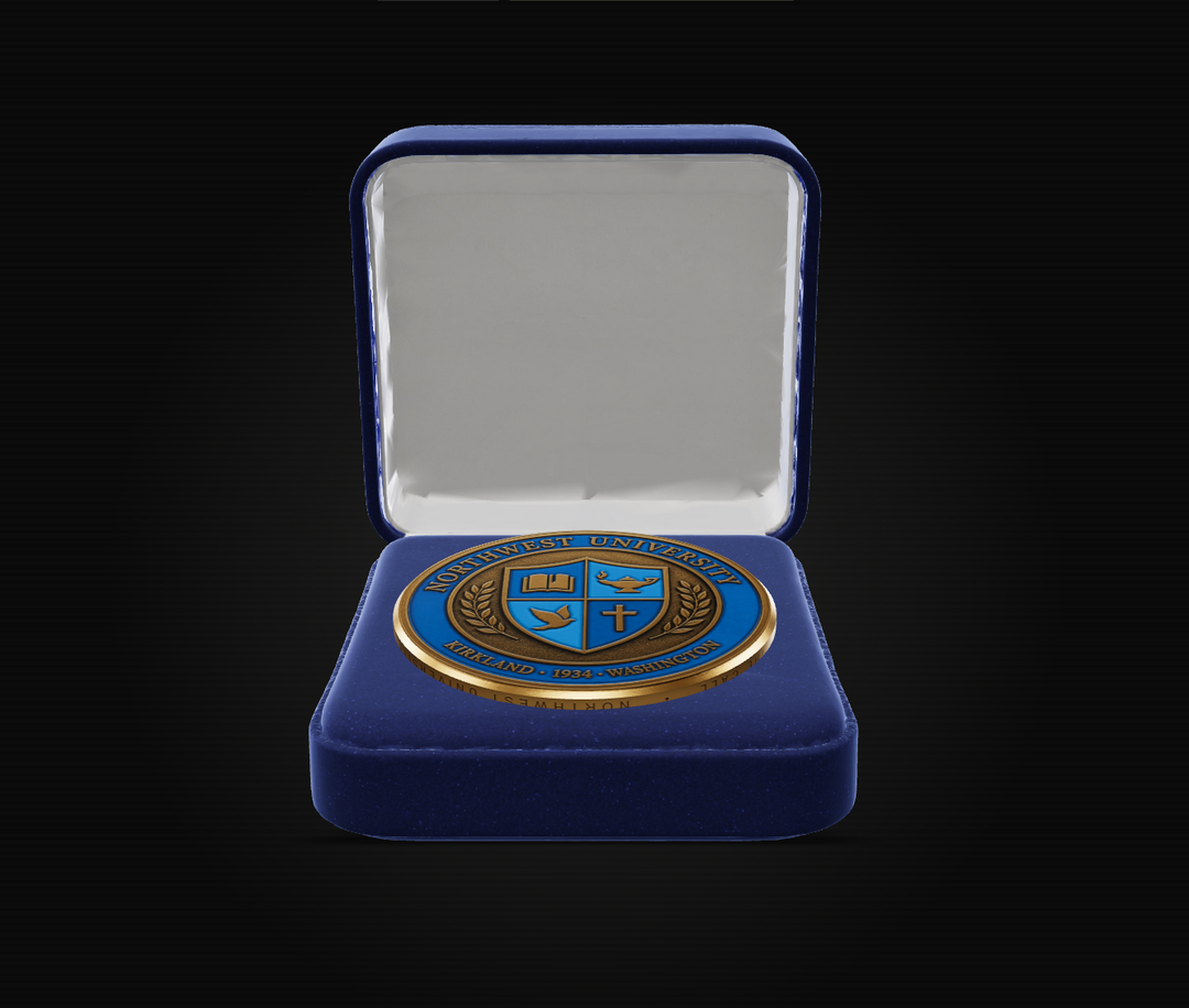An open royal blue velvet presentation box with a white interior, displaying a round blue and gold commemorative coin or medallion on a black background.