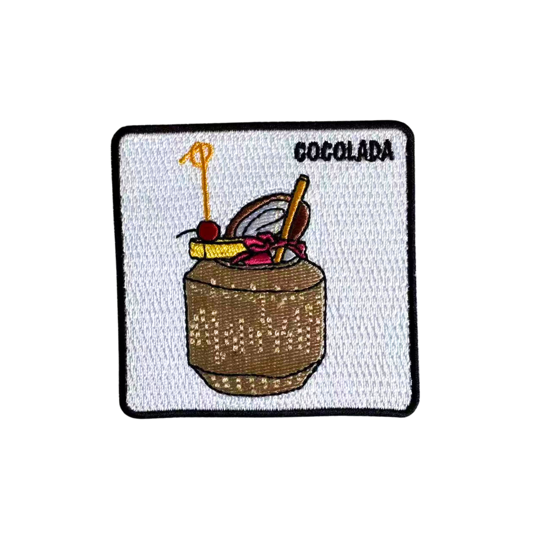 An embroidered square patch featuring a brown coconut-style drink with a yellow straw, cherry, and a red bow, with the word 'COCOLADA' in black at the top right, on a white background with a black border.